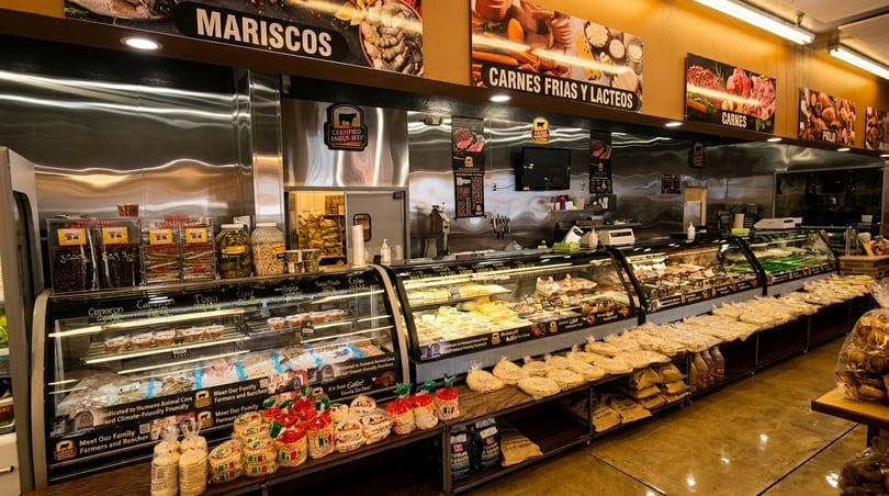 A brightly lit grocery store aisle with refrigerated display cases of meats, cheeses, and baked goods.
