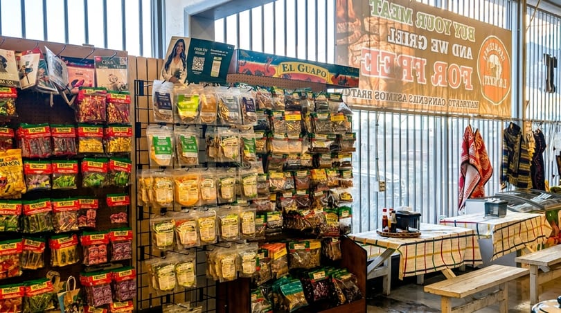 A store interior with shelves of packaged snacks and spices, and tables with checkered cloths.